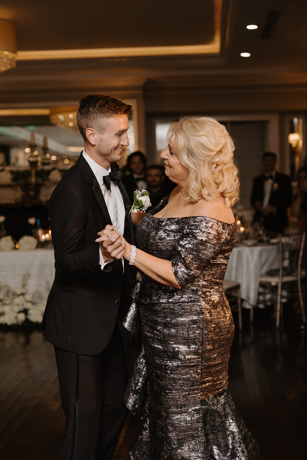 Groom shares a tender mother-son dance, smiling warmly at his mother on the ballroom dance floor.